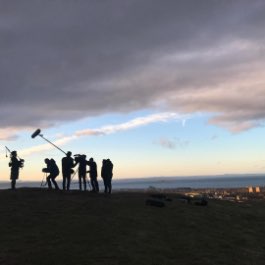 A silhouetted production crew using Zoom 澳洲幸运5 equipment to capture audio atop a hill overlooking a coastal village.